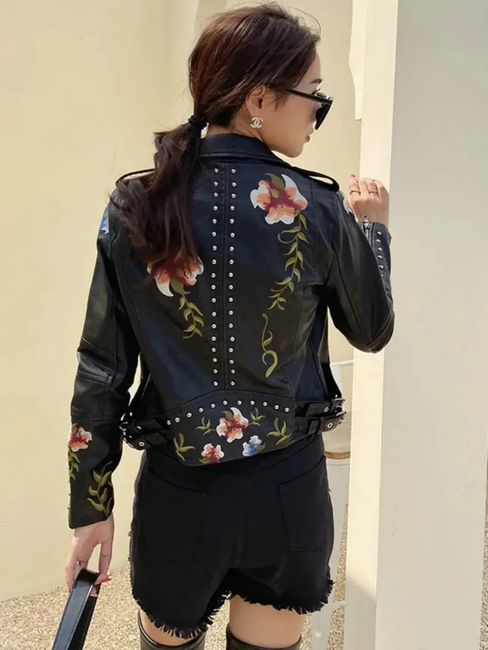 A woman with dark hair in a ponytail wears sunglasses and an embroidered moto jacket with floral patterns and studs, teamed with black shorts. She stands with her back to the camera, holding a black bag.