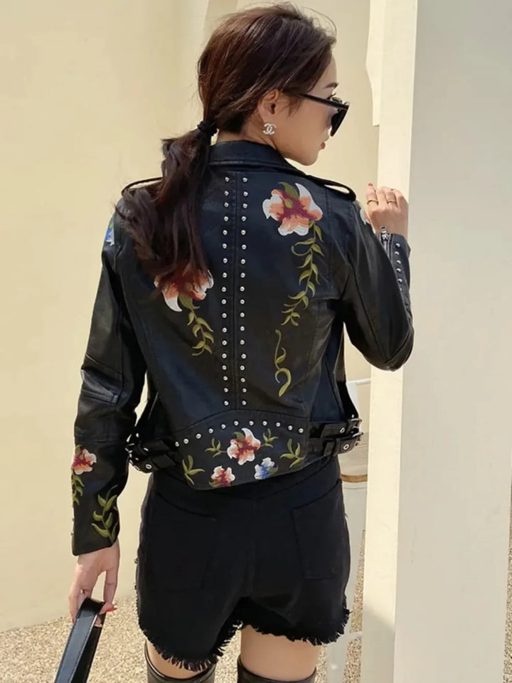 A woman with dark hair in a ponytail wears sunglasses and an embroidered moto jacket with floral patterns and studs, teamed with black shorts. She stands with her back to the camera, holding a black bag.