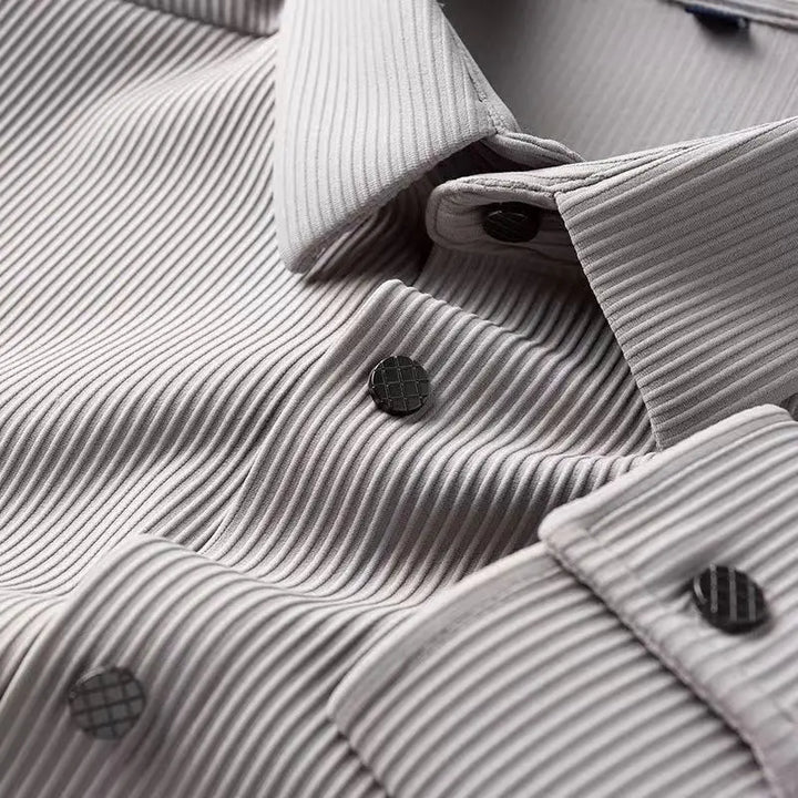Close-up of a light gray, textured dress shirt with vertical ribbed patterns and dark buttons featuring a grid design. Crafted from wrinkle-resistant fabric, the shirt is neatly folded, highlighting the collar and front buttons.