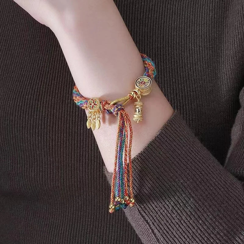 A person wears a multicolored braided bracelet with gold charms and tassels, featuring adjustable Tibetan craftsmanship, on their wrist. The person is dressed in a ribbed, long-sleeve, dark brown top.