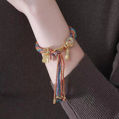 A person wears a multicolored braided bracelet with gold charms and tassels, featuring adjustable Tibetan craftsmanship, on their wrist. The person is dressed in a ribbed, long-sleeve, dark brown top.