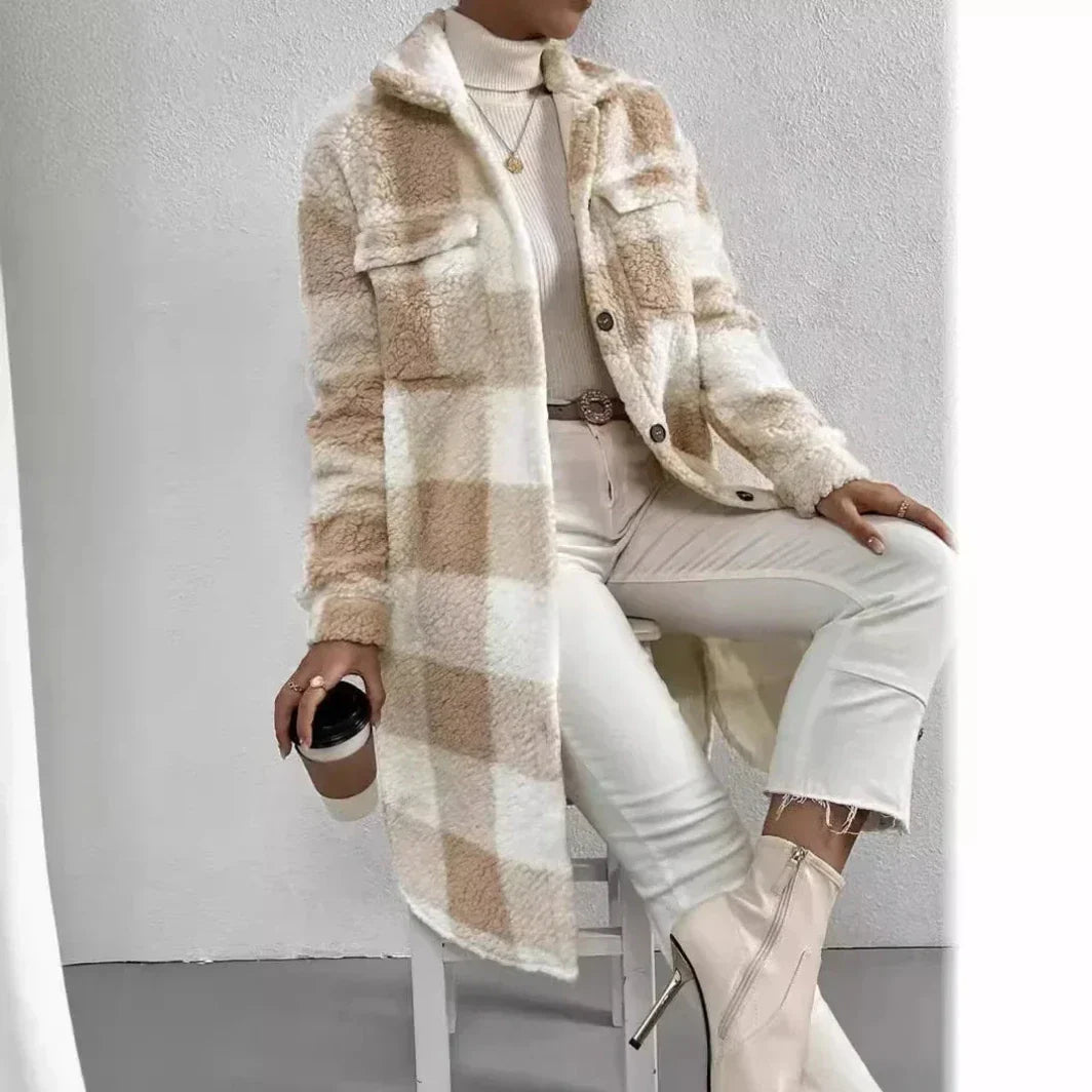 A person sits on a white stool wearing a cream turtleneck, open lapel light beige plaid Sherpa coat, white pants, and heeled ankle boots, holding a takeaway coffee cup. The background is minimal and light-colored.
