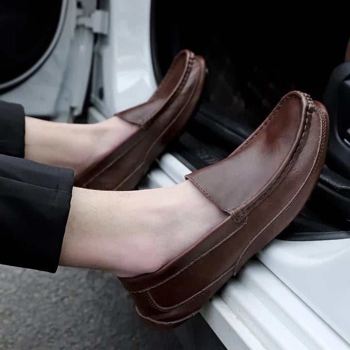 A person wearing genuine leather loafers with black pants sits in the open doorway of a white car, their feet resting on the frame—showcasing the style and comfort of cushioned insoles and hand-stitched moccasin details.