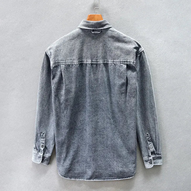 A gray washed denim overshirt made from natural cotton, displayed on a wooden hanger and shown from the back against a plain light gray wall.