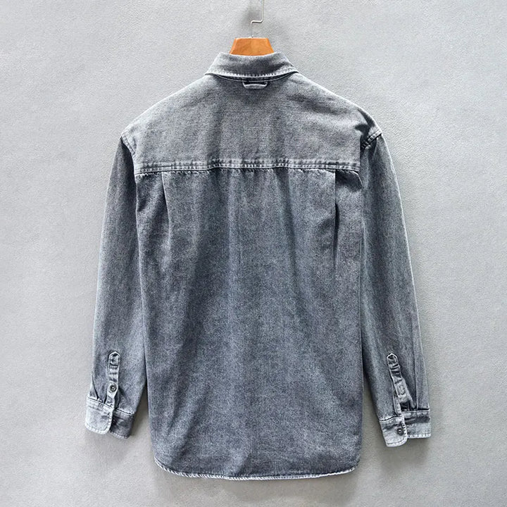 A gray washed denim overshirt made from natural cotton, displayed on a wooden hanger and shown from the back against a plain light gray wall.