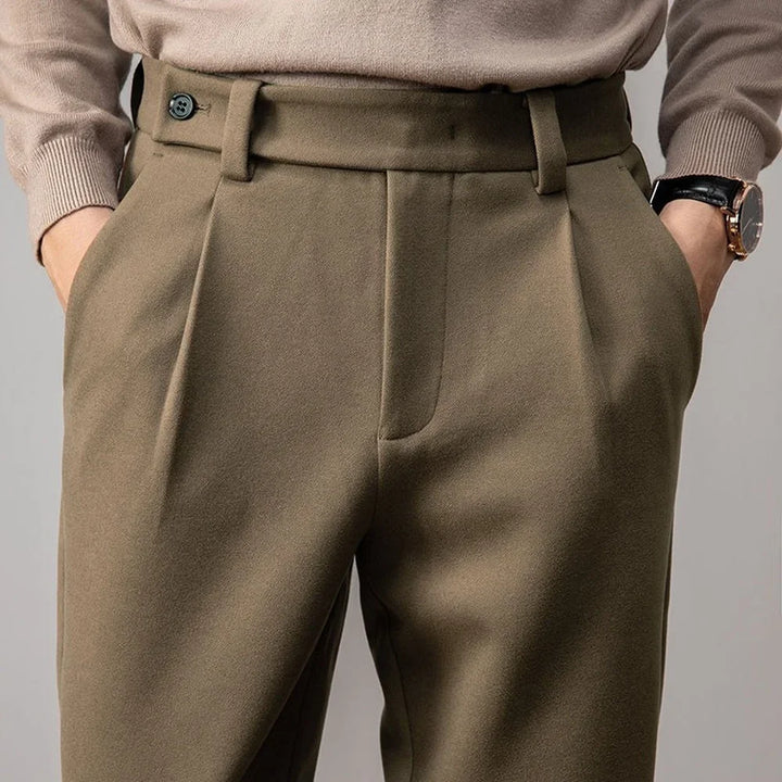 A close-up of a person wearing high-waisted, pleated, olive green tailored slacks with hands in the pockets. The person is also wearing a light beige sweater and a wristwatch.