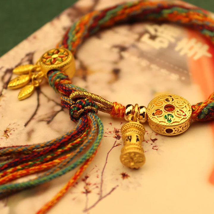 A colorful braided bracelet showcasing Tibetan craftsmanship, with gold charms—a round disc with red and green designs, a cylindrical bead, and small gold leaves—rests on a book adorned with floral artwork and Asian script.