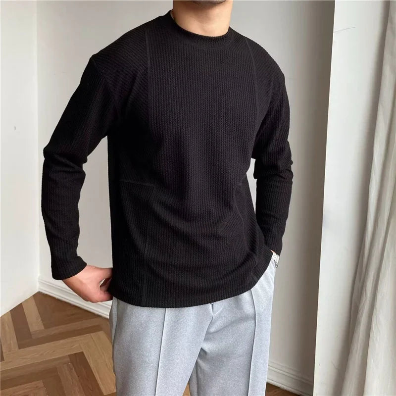 A person stands indoors wearing a black ribbed knit Grayson Crew Classic long-sleeve shirt and light gray pants with hands in pockets. The background features white walls and a wooden floor with a herringbone pattern.