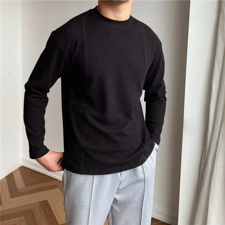 A person stands indoors wearing a black ribbed knit Grayson Crew Classic long-sleeve shirt and light gray pants with hands in pockets. The background features white walls and a wooden floor with a herringbone pattern.