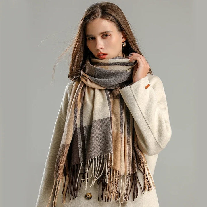 A woman wearing a cream-colored coat and a large, plaid cashmere blend scarf in shades of beige, brown, and gray stands against a plain gray background, looking at the camera with a neutral expression.