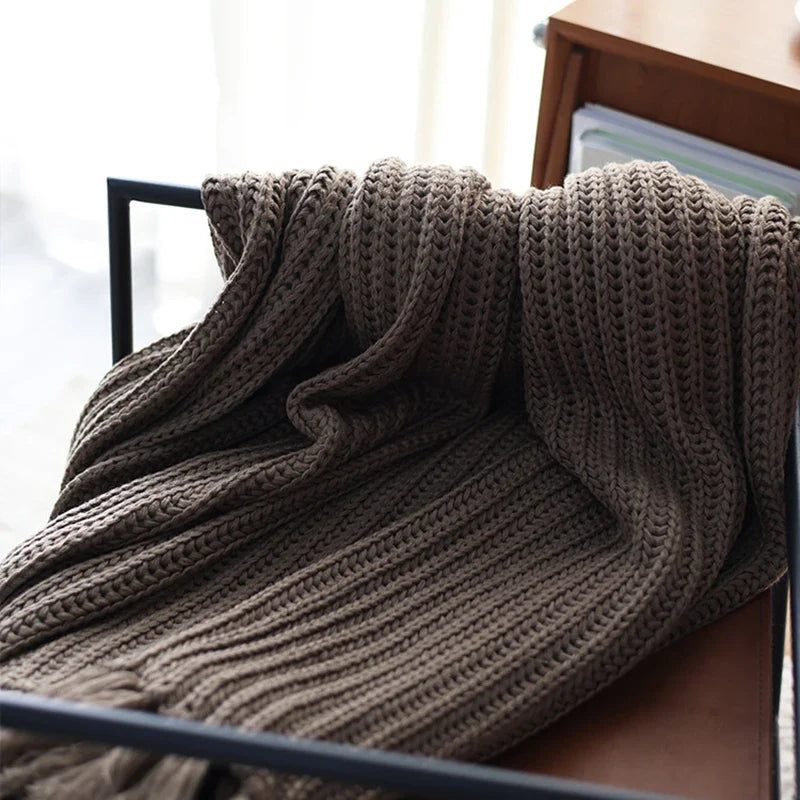 A chunky weave throw in brown is draped over the back of a black metal chair, with a wooden shelf and books visible in the background. Soft daylight filters in from the left side, highlighting this cozy, stress reducing blanket.