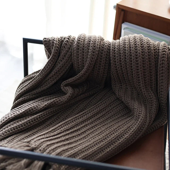 A chunky weave throw in brown is draped over the back of a black metal chair, with a wooden shelf and books visible in the background. Soft daylight filters in from the left side, highlighting this cozy, stress reducing blanket.