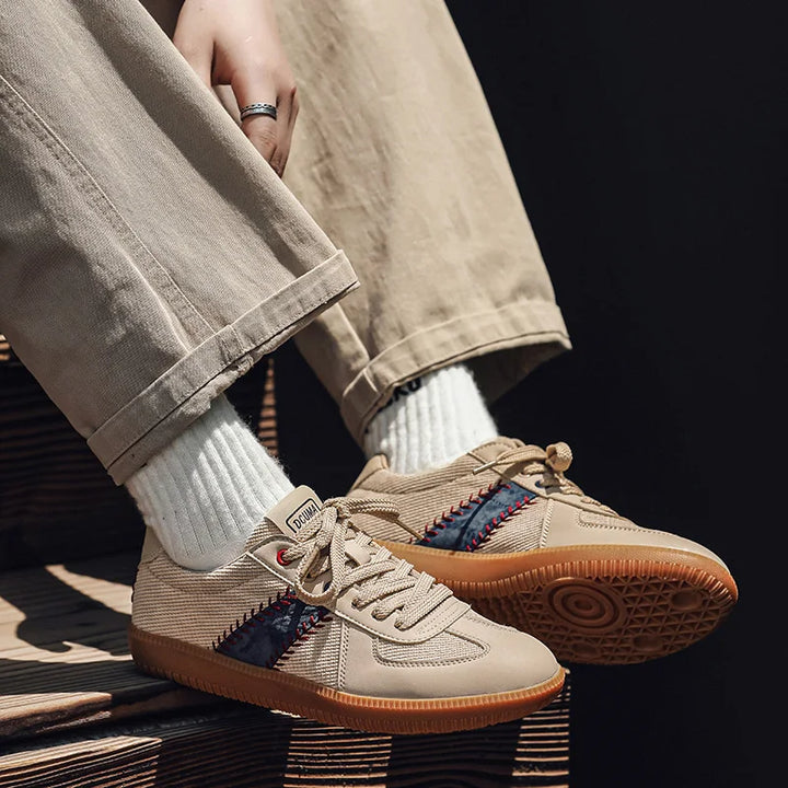 A person wearing beige pants and white socks sits on wooden steps, showing off baseball inspired shoes with a brown sole, navy and red side accents, detailed stitching, and lightweight breathable sneakers design.