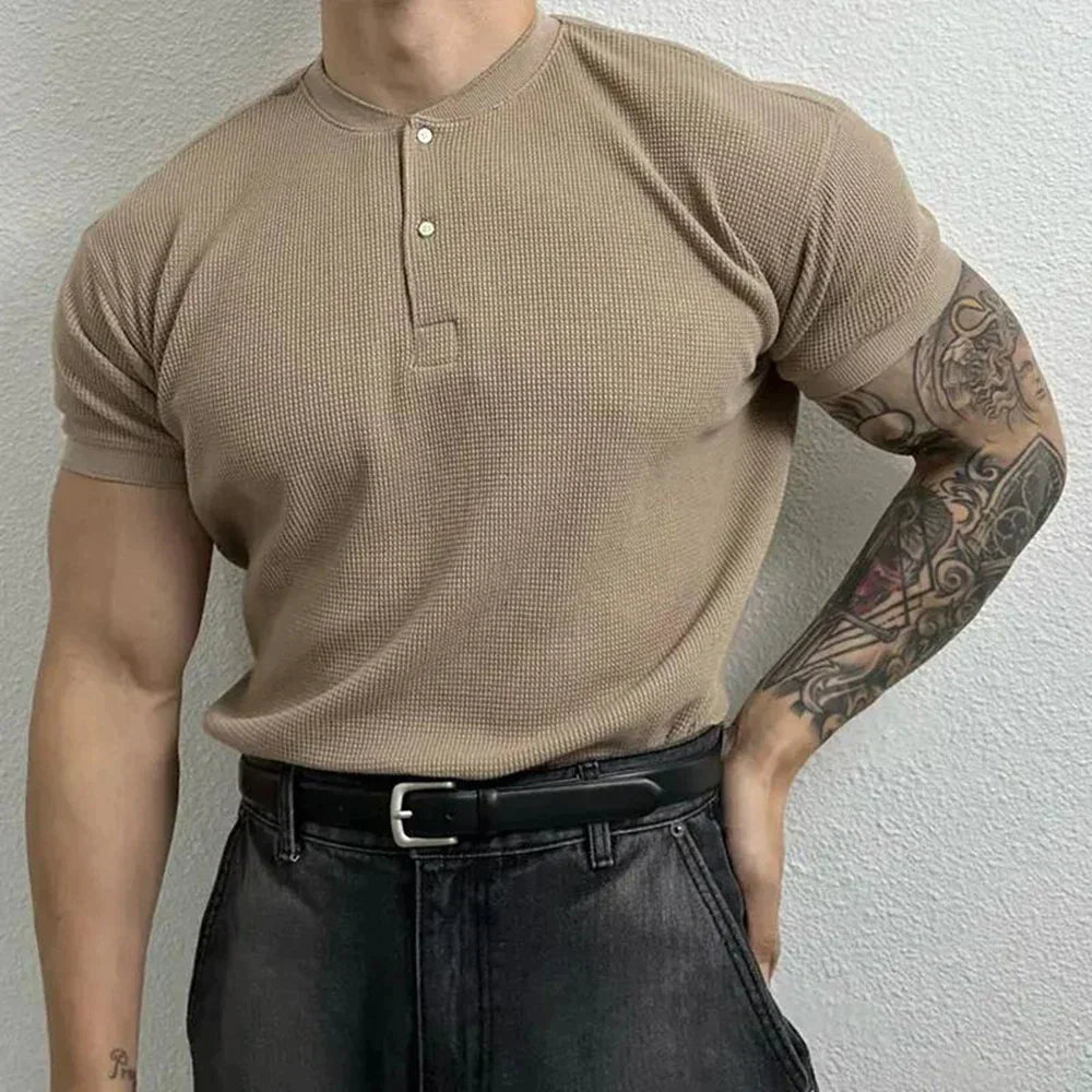 A muscular person wearing a ribbed Henley, black belt, and dark jeans stands against a light-colored textured wall, showing a tattooed right arm. The form-fitting shirt highlights their physique.
