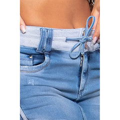 Close-up of a person wearing light blue jeans with a high waist, featuring an elastic, ribbed waistband and a decorative denim tie at the front. The person has light-colored nail polish and is holding the waistband.