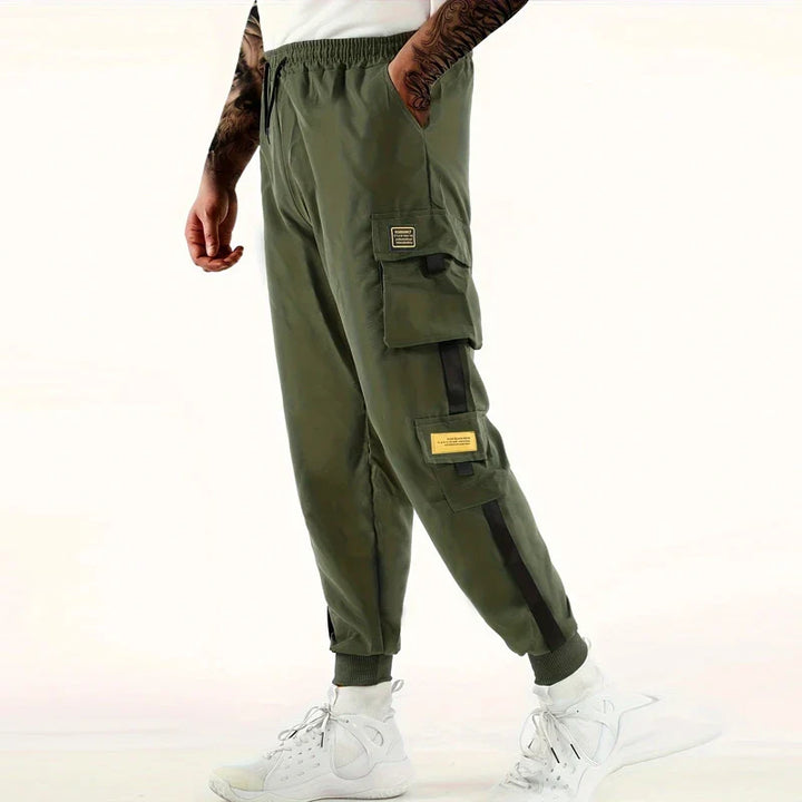 A person wearing olive green cargo pants with an elastic waist, black and yellow patches, ribbed ankle cuffs, white athletic shoes, and a white background. Only the lower half of the body is visible.