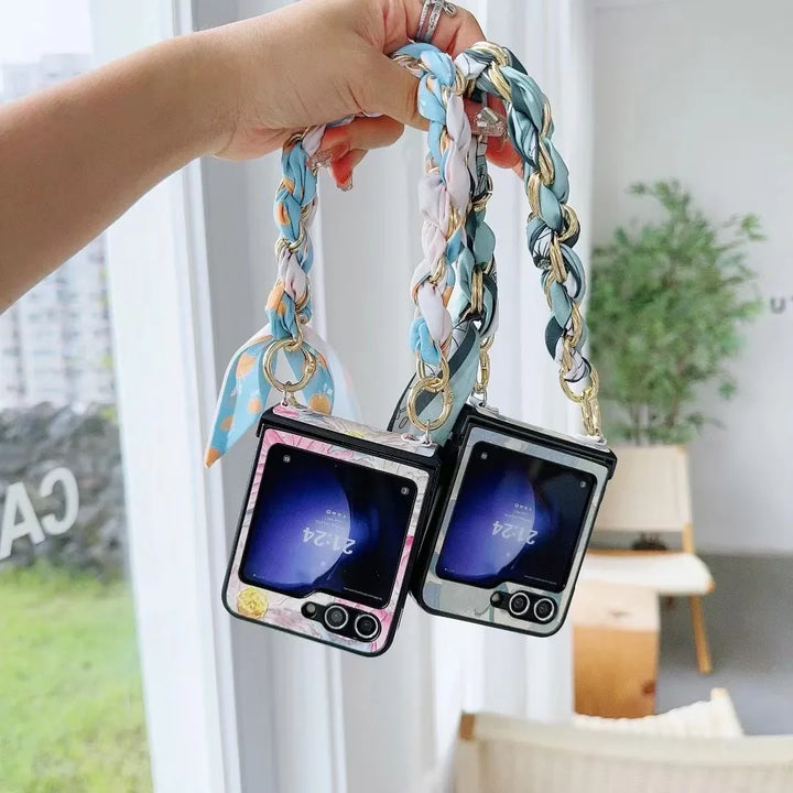 A hand holds two foldable smartphones with colorful braided chain straps—one pink and one black—offering slim design in a bright room with large windows and white furniture.