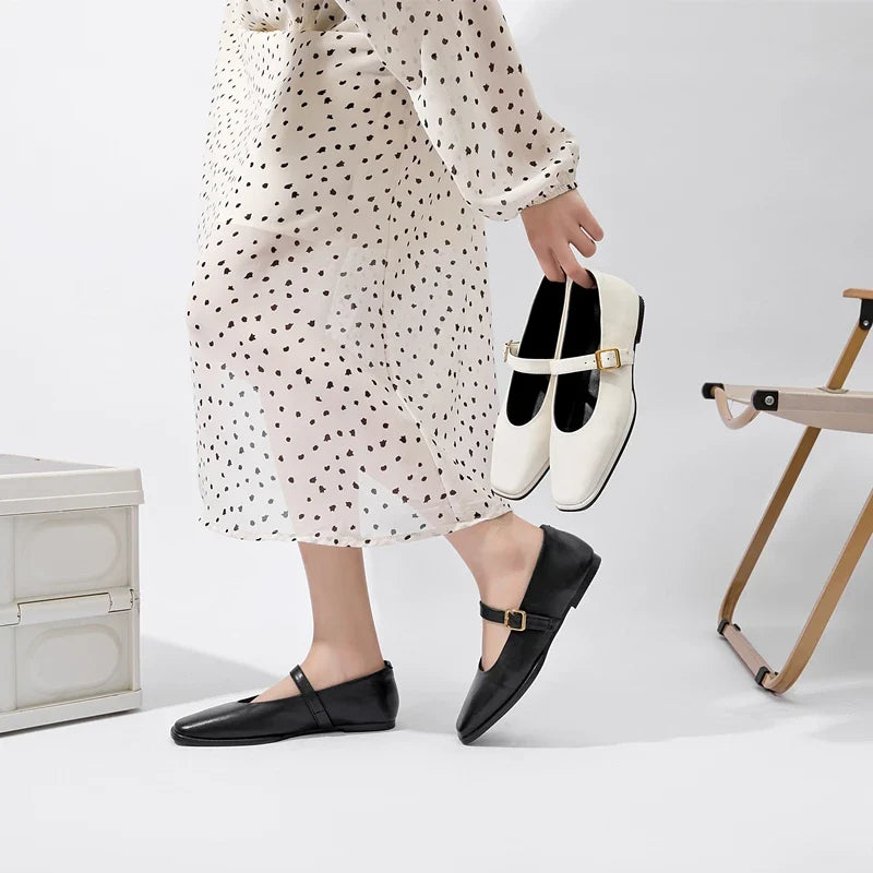 A person in a white dress with black polka dots wears black Mary Jane shoes and holds a pair of Augusta Leather Ballet Flats in one hand, standing near a folding chair and a white storage box.