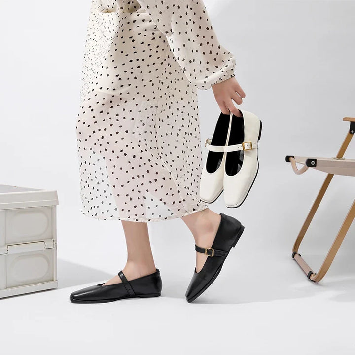 A person in a white dress with black polka dots wears black Mary Jane shoes and holds a pair of Augusta Leather Ballet Flats in one hand, standing near a folding chair and a white storage box.