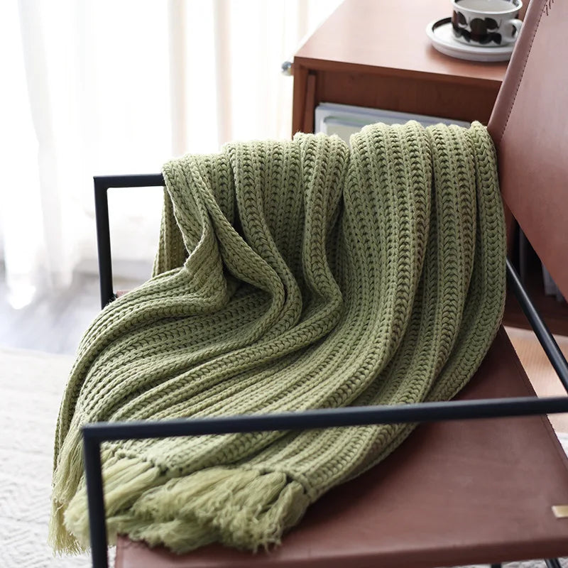 A light green earth-tone throw with fringe is draped over a brown chair next to a small wooden table with a cup and saucer. Sunlight filters through sheer curtains in the background, creating a calming, stress-reducing blanket setting.