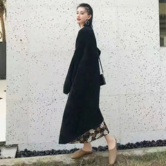 A woman stands outdoors against a speckled white wall, wearing a long black coat with a relaxed drape fit over a floral dress, beige shoes, and carrying a black purse on her shoulder.