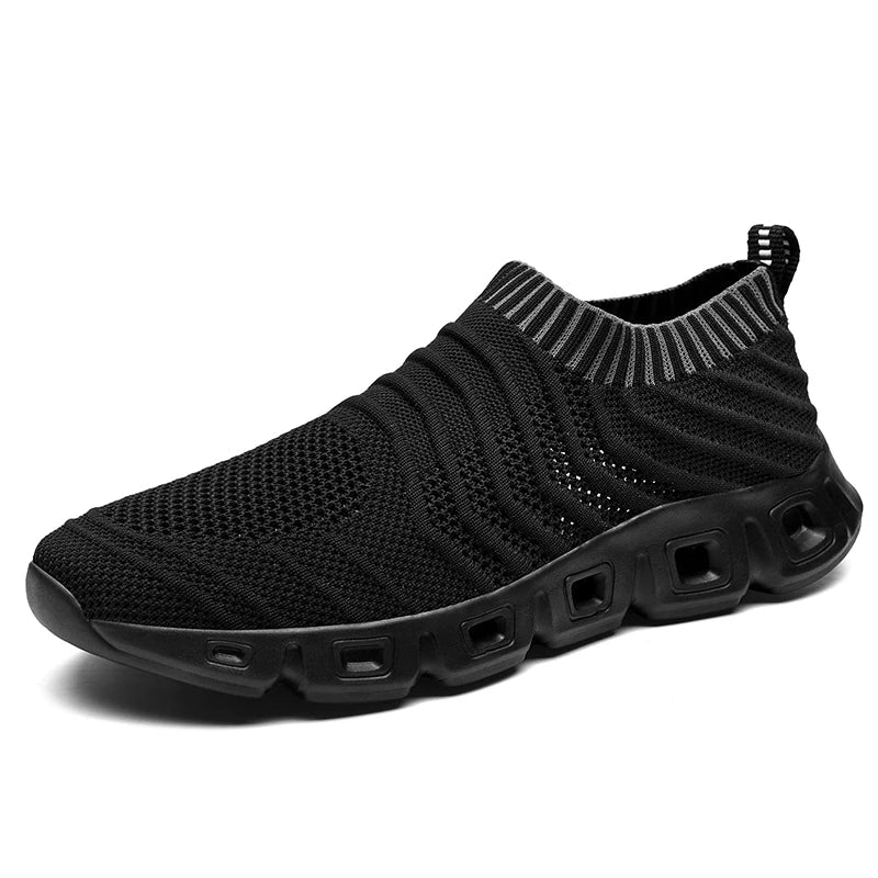 Black slip-on athletic shoe with a knit mesh upper, ribbed detailing, and a thick cushioned sole featuring rectangular cutouts. The shoe offers a sock-like fit with a pull tab at the heel and a low ankle collar with gray striped accents.