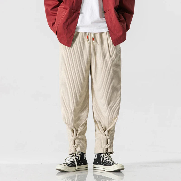 Person wearing a red jacket, white shirt, beige cotton blend sweatpants with an adjustable fit and drawstring waistband, tied cuffs, and black high-top sneakers stands against a plain white background.