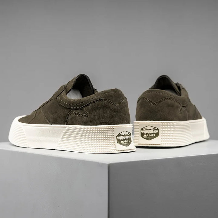A pair of dark olive-green suede Lace-Up Sneakers with thick white textured soles are displayed on a gray platform. The back heel features a rectangular gold and black “JIAMEI” label with a mountain logo. Comfortable cushioning adds extra support.
