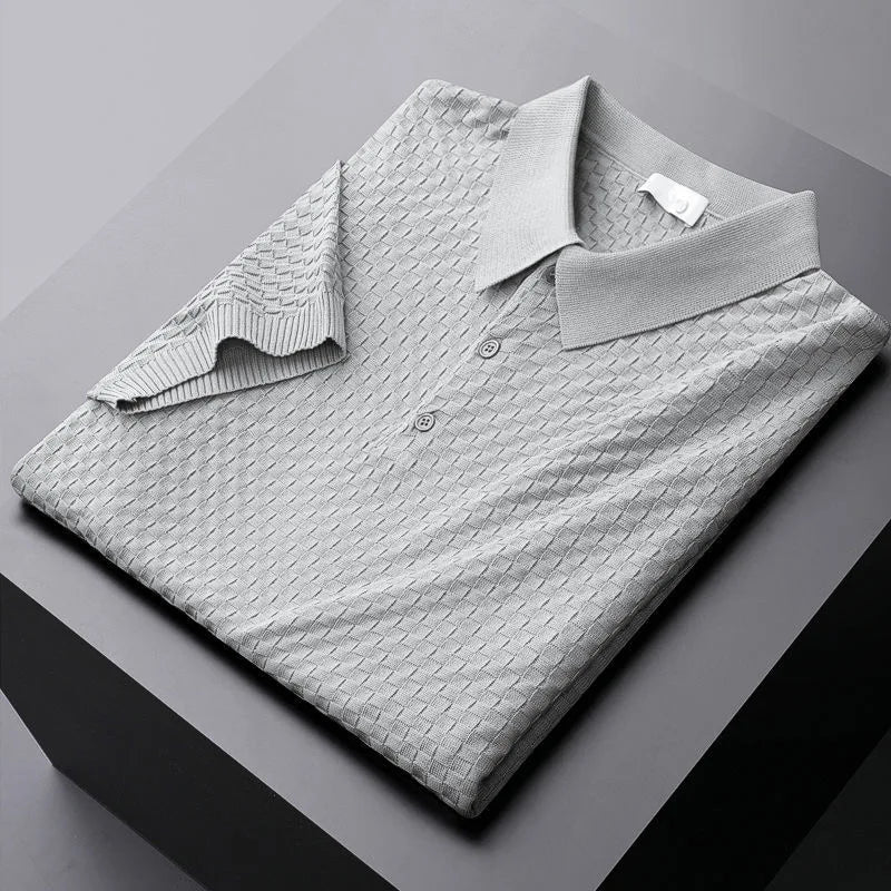 A neatly folded light gray waffle jacquard polo shirt with a collar and two buttons, placed on a dark gray surface. Part of the left sleeve is turned up to show the unique inner fabric pattern.