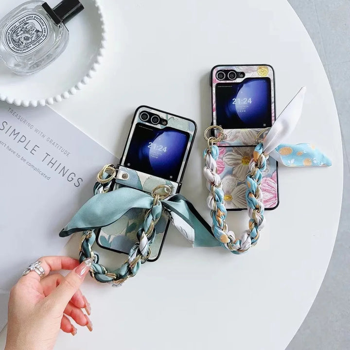 Two smartphones with slim design and chic chain straps rest on floral cases beside a glass perfume bottle and a book, as a hand reaches for one.