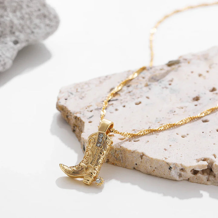 A gold cowgirl necklace with a detailed cowboy boot pendant, featuring decorative engravings and a small gemstone, displayed on a textured stone surface—a perfect piece of western jewelry.