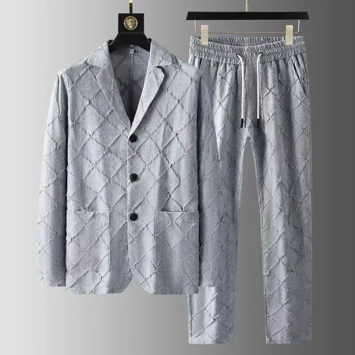 A light grey luxury two-piece set featuring a buttoned blazer with two pockets and matching relaxed fit trousers, both adorned with a quilted diamond pattern and hanging on black hangers against a grey gradient background.