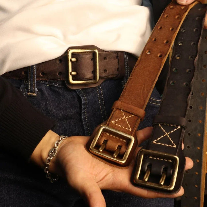 A person in jeans and a white shirt holds two cowhide belts, one brown and one black, both with large brass buckles and visible stitching. Another brown belt with a rugged aesthetic is worn around their waist.