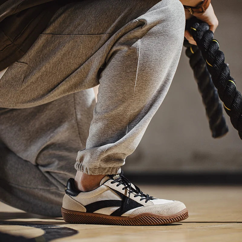 Person in grey sweatpants kneeling on a gym floor, holding a thick workout rope. The focus is on their Olympism 1984 Pro Sneakers with beige and black uppers, brown soles, and EVA midsole cushioning inspired by 80s aesthetics.