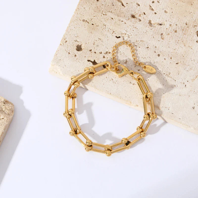 A gold statement chain bracelet with small gold beads sits on a textured beige stone surface, casting a subtle shadow. The bracelet features an adjustable clasp and a small tag attached.