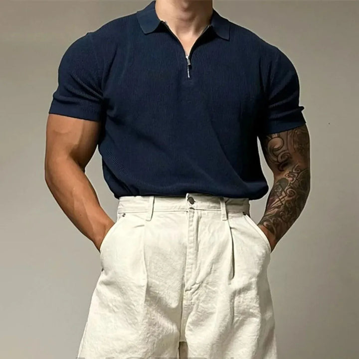 A person with a muscular build wears a navy quarter zip polo with ribbed detailing and high-waisted white pleated pants, hands in pockets. One tattooed forearm is visible. The background is plain and neutral.