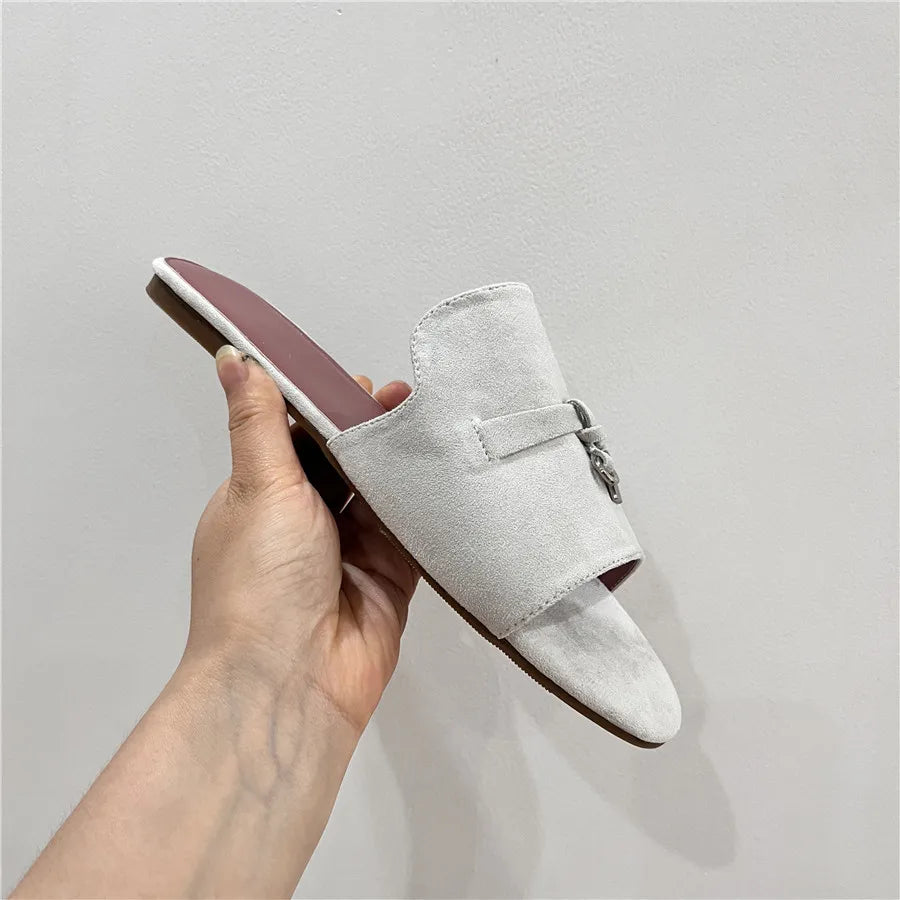 A hand holds a minimalist white suede finish slip-on sandal with an open toe, a flat sole, and a small decorative metallic accent on the upper. The background is a plain light-colored wall.