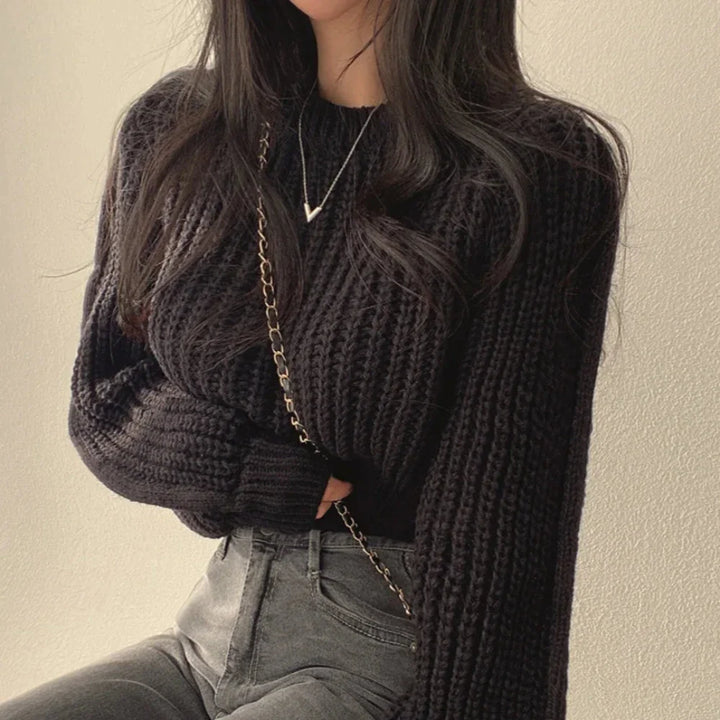 A woman wearing a chunky black sweater in soft knit material, high-waisted gray jeans, a gold chain purse, and a delicate pendant necklace poses against a light-colored wall. Her long dark hair is loose over her shoulders.