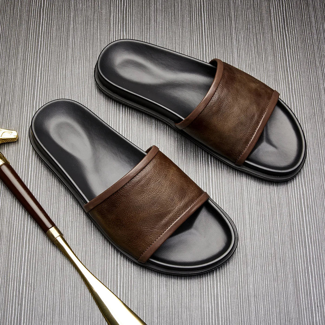 A pair of black and brown handmade leather sandals are placed on a textured gray surface next to a gold and brown shoehorn.
