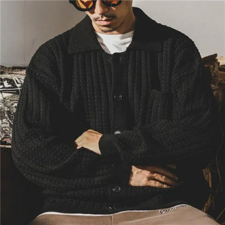 A man wearing orange sunglasses, a white t-shirt, and a chunky knit black button-up wool cardigan sits with his arms crossed on his lap, partially facing the camera.