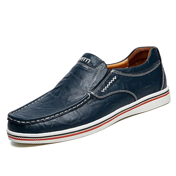 A dark blue leather slip-on shoe with white stitching, a white sole featuring a red stripe, and a tan interior lining. These boat-style loafers have a casual, moccasin-inspired design.