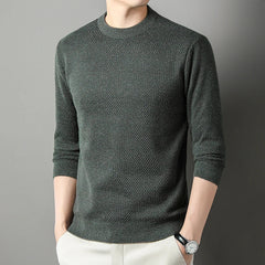 A person wearing a versatile, dark green high-quality cotton sweater with light-colored pants stands with hands in pockets against a plain gray background. The relaxed fit sweater adds comfort, while the person’s head is not fully visible.