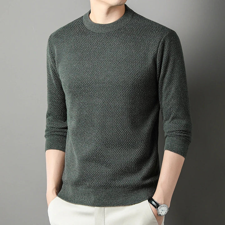 A person wearing a versatile, dark green high-quality cotton sweater with light-colored pants stands with hands in pockets against a plain gray background. The relaxed fit sweater adds comfort, while the person’s head is not fully visible.