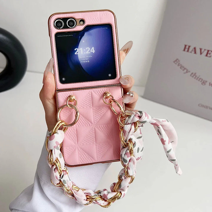 A hand holds a pink, quilted Samsung Galaxy Z Flip with a decorative gold chain and floral scarf attached. The phone features shockproof protection, and its small external screen displays the time, 21:24. A blurred background includes a sign with partial text.