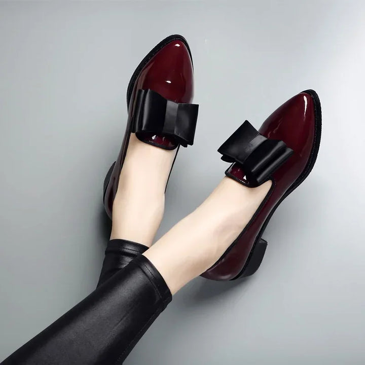 A person wearing shiny burgundy patent leather loafers with a statement bow and low block heel, paired with black faux leather leggings, against a plain gray background.