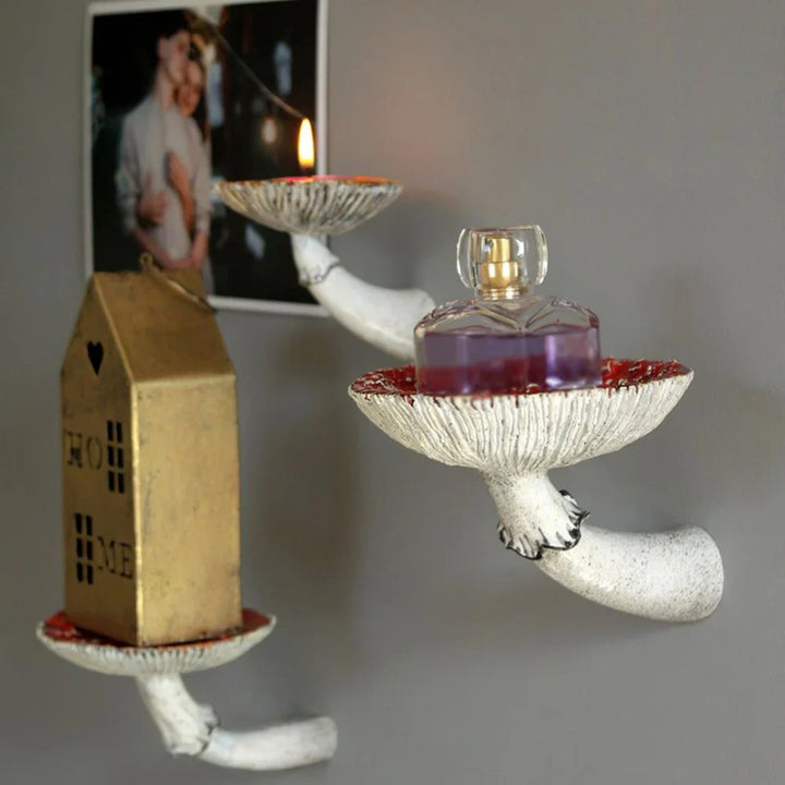 Two wall-mounted mushroom floating shelves shaped like white mushrooms display unique home decor: a gold house-shaped ornament, a purple perfume bottle, and a lit candle. A photo of a couple is pinned to the gray wall behind them.