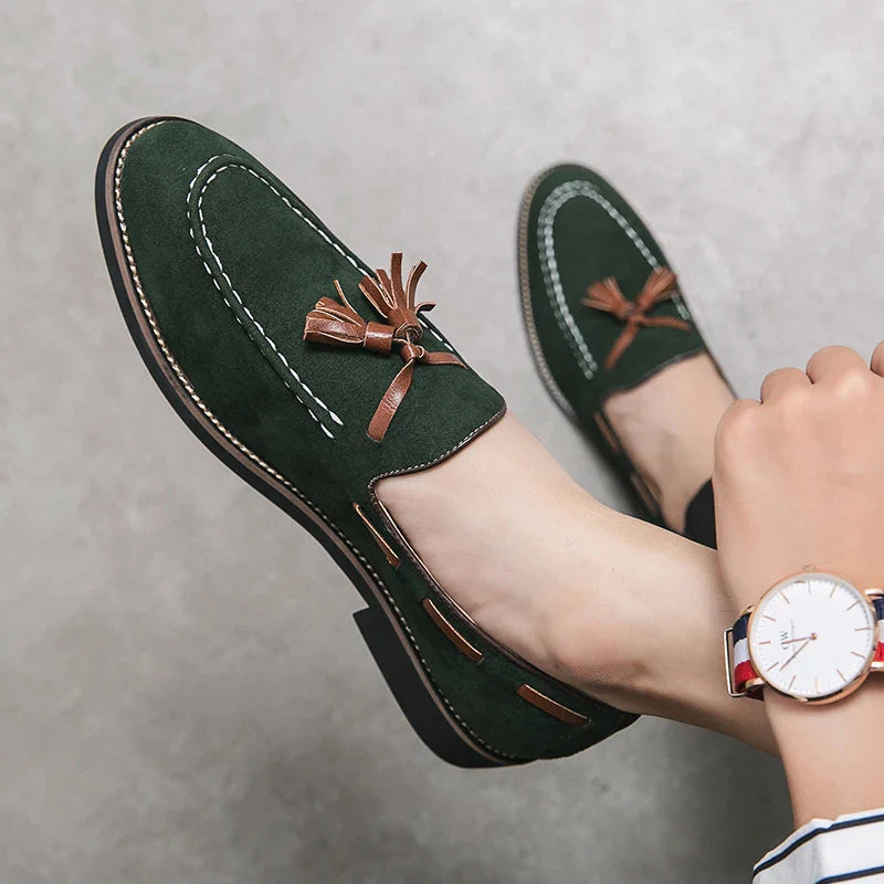 A person wearing dark green tassel loafers with brown accents, durable construction, and white stitching, paired with a wristwatch and striped pants, rests their feet against a light gray background.