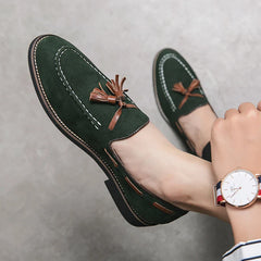 A person wearing dark green tassel loafers with brown accents, durable construction, and white stitching, paired with a wristwatch and striped pants, rests their feet against a light gray background.
