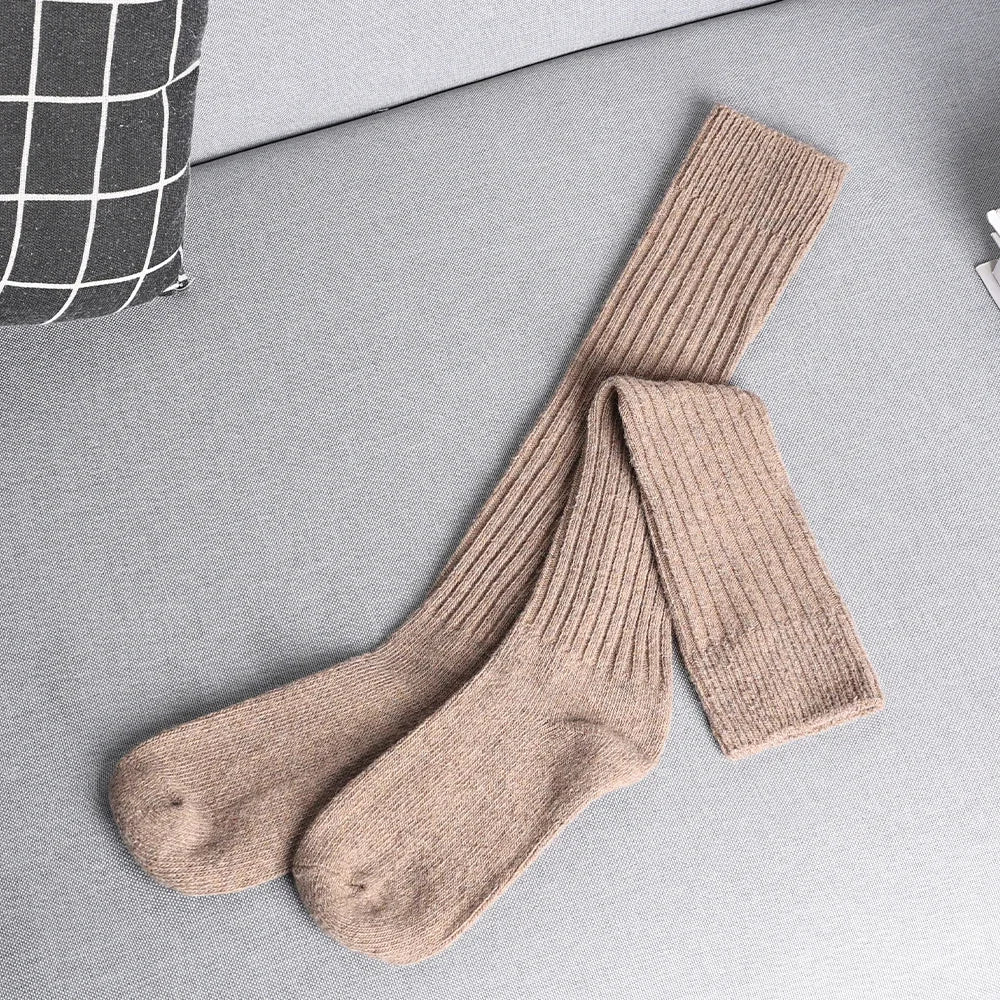 A pair of long, ribbed, beige knit socks crafted from a Cashmere cotton blend, laid out on a light grey sofa next to a black-and-white grid-patterned pillow.
