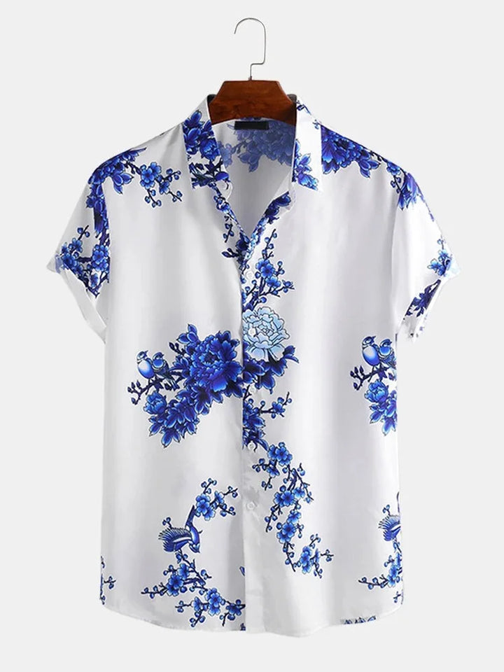 A summer-ready, short-sleeved white button-up shirt featuring a blue floral and branch pattern, crafted from a soft cotton-silk blend and displayed on a wooden hanger against a plain light background.